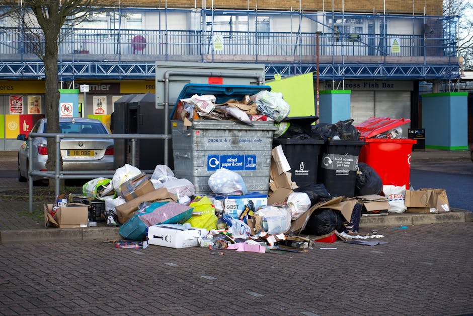 A collection of overflowing rubbish bins and scattered waste is positioned on a paved area beside a parking space, with a silver car parked adjacent to the bins. The waste includes black and red plast