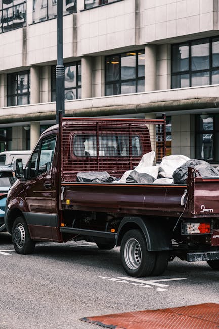 A small dark red flatbed truck parked on an urban street in front of a modern, multi-storey office building featuring large glass windows and a light-colored façade. The truck's cargo area is filled